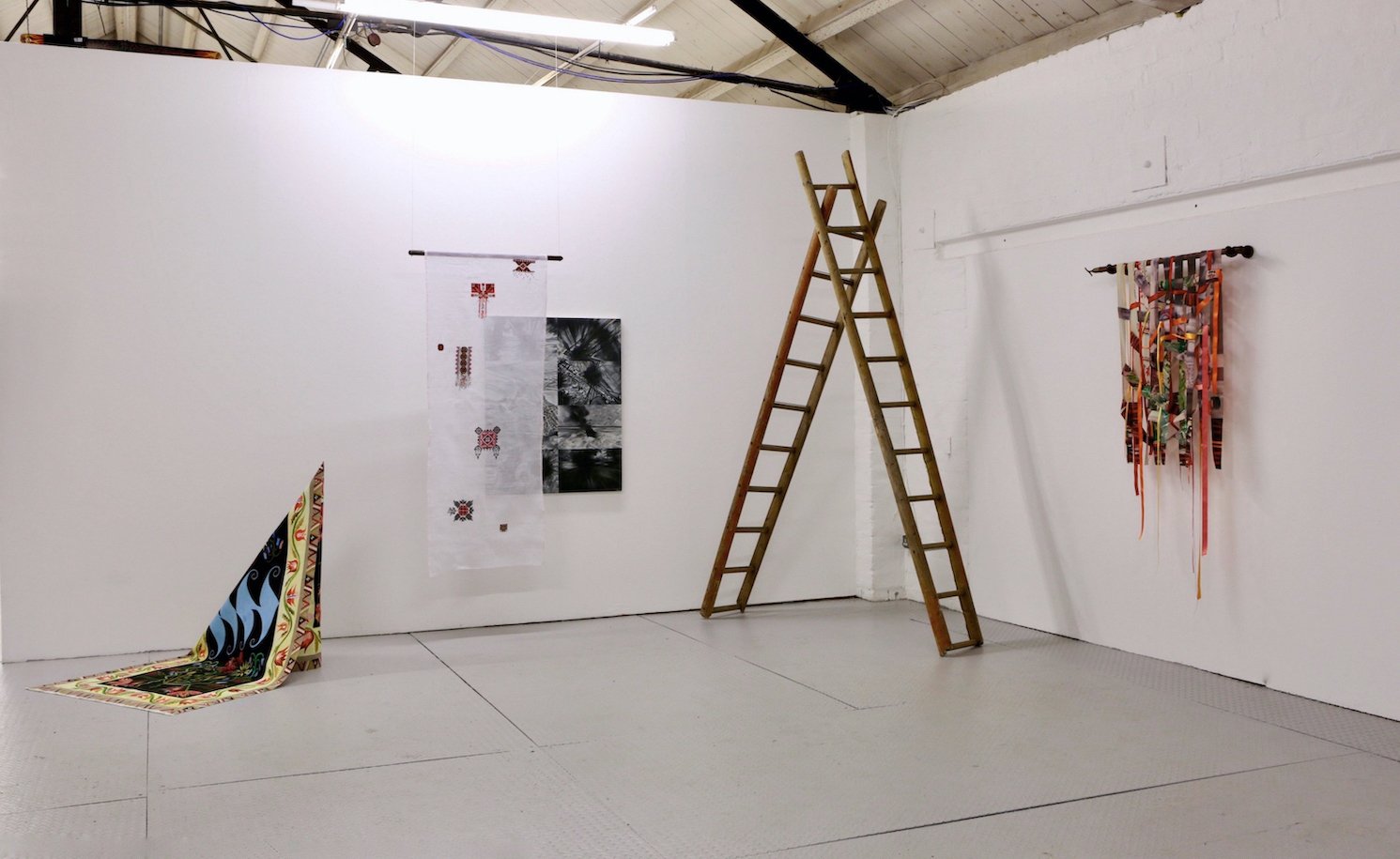 An installation with paintings, textiles and ladders is shown as a physical collage. The space they sit within is white and lit from above.