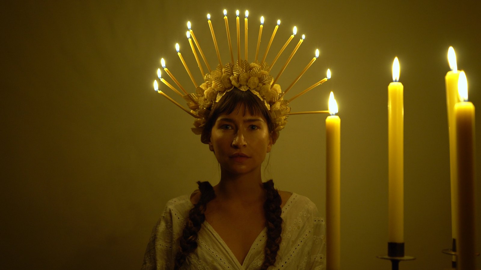 A woman wearing a wreath made of thin beeswax candles is lit. She stares directly into the camera. Hanging from the wreath are severed braids.