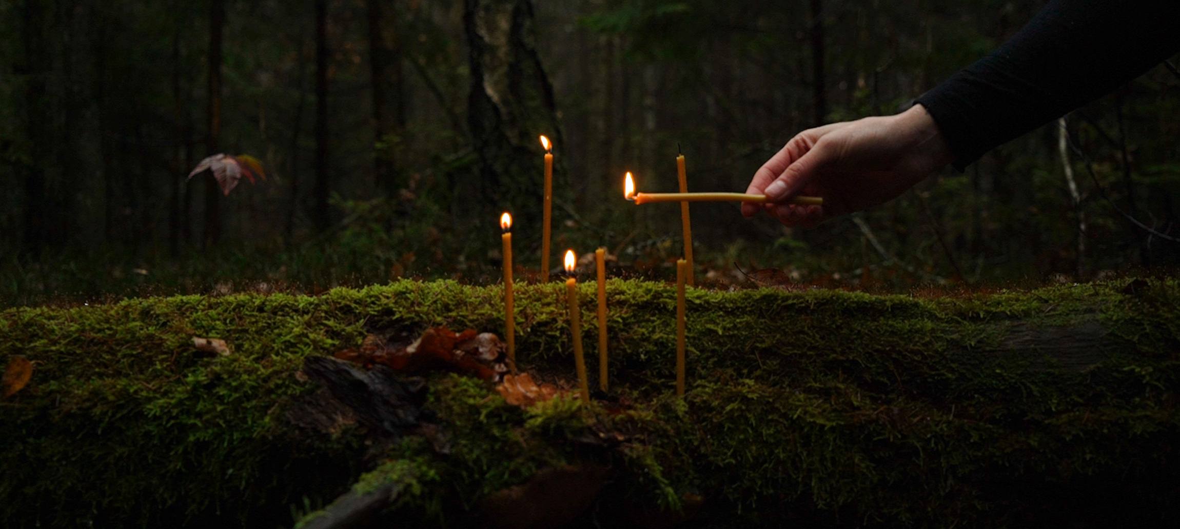 A woman wearing a wreath made of thin beeswax candles is lit. She stares directly into the camera. Hanging from the wreath are severed braids.